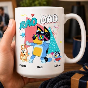 May include: A white ceramic mug featuring a colorful graphic of three cartoon dogs in a dabbing pose. The text RAD DAD is printed at the top, with the names EMMA, DAD, and LIAM printed below each respective character. The design includes geometric shapes.