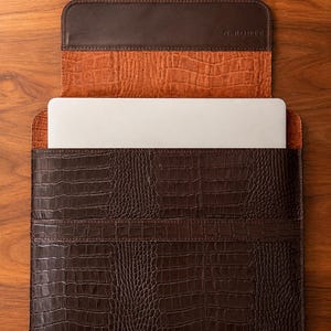 May include: A brown leather laptop sleeve with a crocodile texture. The sleeve is open, revealing a white laptop inside. The interior is a lighter shade of brown. The top flap has the text "N. ROUTE".