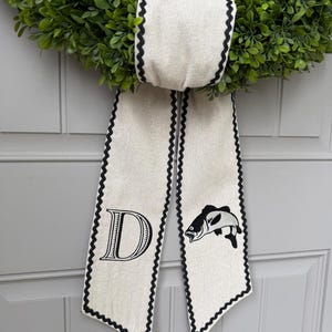 May include: A green boxwood wreath with a cream-colored bow. The bow has a black scalloped edge and features a large letter "D" and a fish illustration. The wreath is hanging on a gray door.