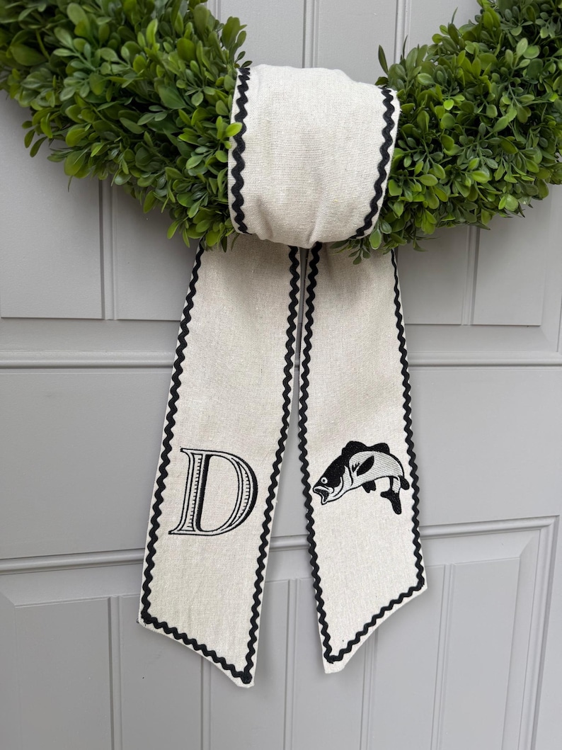 May include: A green boxwood wreath with a cream-colored bow. The bow has a black scalloped edge and features a large letter "D" and a fish illustration. The wreath is hanging on a gray door.