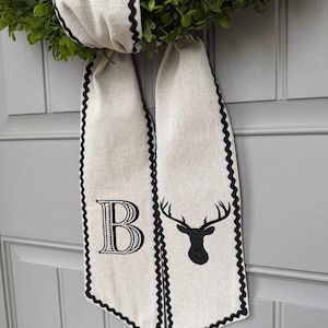 May include: A decorative wreath with a green, leafy base and a cream-colored ribbon. The ribbon has a black scalloped edge and features a large letter "B" and a black deer head silhouette.