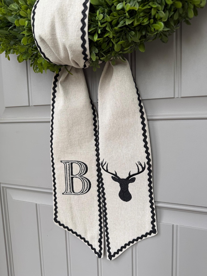 May include: A decorative wreath with a green, leafy base and a cream-colored ribbon. The ribbon has a black scalloped edge and features a large letter "B" and a black deer head silhouette.