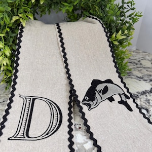 May include: Two beige linen towels with black scalloped edges. One towel features a large black letter "D", the other has a black and gray fish graphic. The towels are displayed against a backdrop of green foliage.
