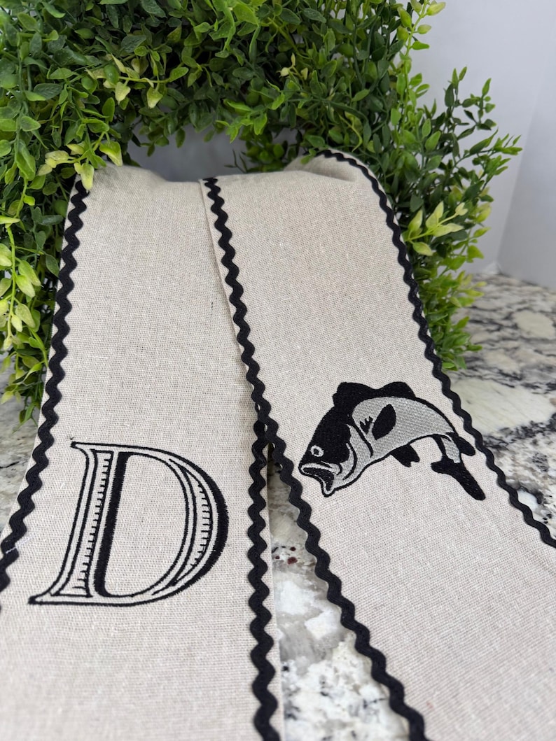 May include: Two beige linen towels with black scalloped edges. One towel features a large black letter "D", the other has a black and gray fish graphic. The towels are displayed against a backdrop of green foliage.