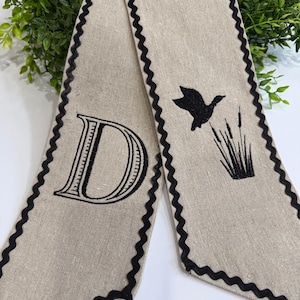 May include: A beige linen scarf with a black scalloped edge. One end features a large letter "D" in black, while the other has a black silhouette of a flying duck and cattails.