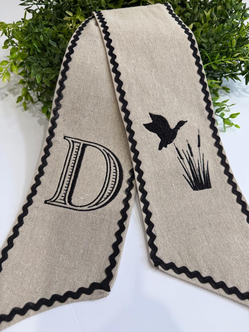 May include: A beige linen scarf with a black scalloped edge. One end features a large letter "D" in black, while the other has a black silhouette of a flying duck and cattails.