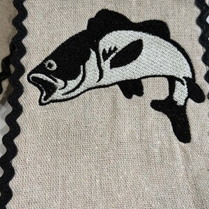 May include: A beige linen fabric with a black and white embroidered fish design. The fish is stylized with open mouth and detailed fins. The edges are trimmed with a black, wavy trim. This item is suitable for kitchen decor.