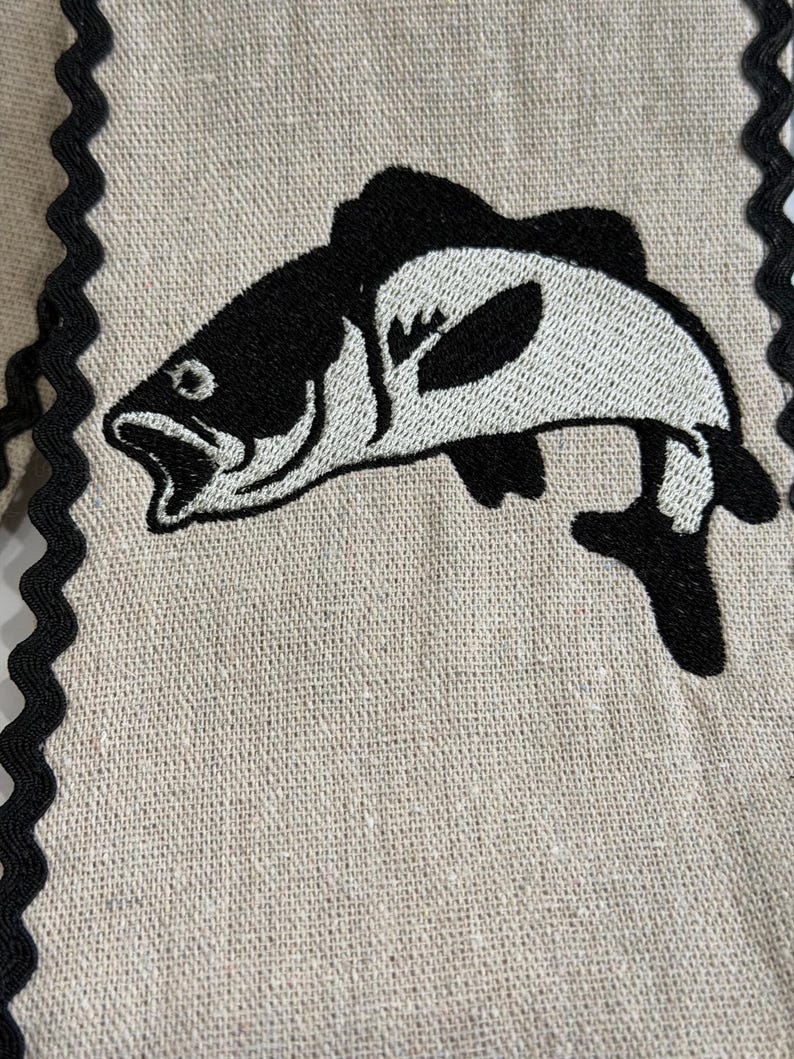 May include: A beige linen fabric with a black and white embroidered fish design. The fish is stylized with open mouth and detailed fins. The edges are trimmed with a black, wavy trim. This item is suitable for kitchen decor.