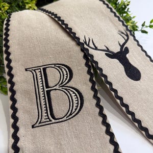May include: Two beige linen decorative pieces with black scalloped edges. One features a large embroidered black letter "B", the other a black silhouette of a deer head with antlers. Green foliage is visible in the background.