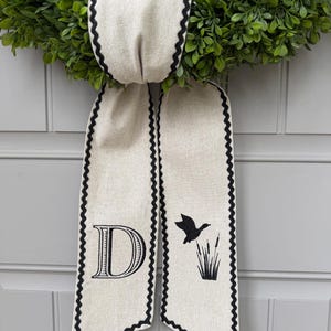 May include: A green wreath with a cream-colored ribbon featuring a black scalloped edge. The ribbon has a large letter "D" and a silhouette of a flying duck with cattails. The wreath is likely for home decor.
