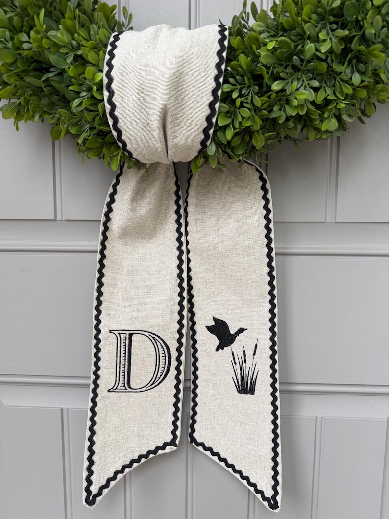 May include: A green wreath with a cream-colored ribbon featuring a black scalloped edge. The ribbon has a large letter "D" and a silhouette of a flying duck with cattails. The wreath is likely for home decor.