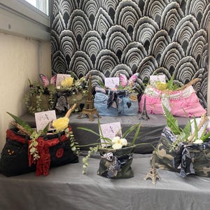 Peut inclure: Présentation de jardinières faites à partir de jeans et de tissus recyclés, remplies de plantes et d'éléments décoratifs. Les jardinières sont fabriquées à partir de jeans et d'autres tissus de différentes couleurs, dont le noir, le bleu, le rose et le camouflage. Chaque jardinière est ornée de nœuds, de papillons et de figurines miniatures de la Tour Eiffel.