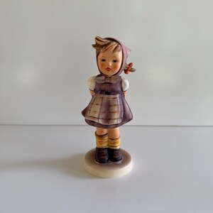 Könnte beinhalten: Eine Keramikfigur einer jungen Person mit einem dunkelvioletten Kopftuch, einem passenden violetten Kleid mit weißem Karomuster, weißen kurzen Ärmeln, gelben Socken und dunkelbraunen Schuhen, die auf einem runden, cremefarbenen Sockel steht.