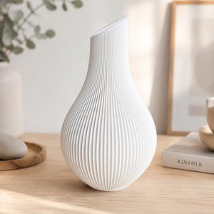 Puede incluir: Un jarrón blanco con una base redondeada y bulbosa que se estrecha hacia una abertura estrecha e inclinada. La superficie exterior presenta una textura acanalada vertical constante que va desde la base hasta el borde superior, creando líneas paralelas.