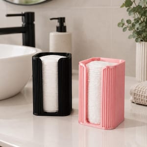 Könnte beinhalten: Zwei rechteckige Wattepadspender, einer in Schwarz und einer in Pink, mit einer vertikal gerippten Textur an der Außenseite. Jeder Spender enthält einen Stapel runder weißer Wattepads, die durch eine U-förmige Aussparung zugänglich sind.