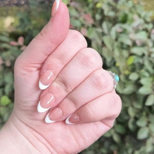 May include: Close-up of a hand with French tip acrylic nails. The nails are a natural pink with white tips. A silver ring with a turquoise stone is visible on the ring finger.