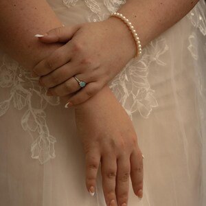 May include: A close-up of hands adorned with a pearl bracelet and a ring featuring a light blue gemstone. The nails are painted with a French manicure. The hands are resting on a white, embroidered fabric.