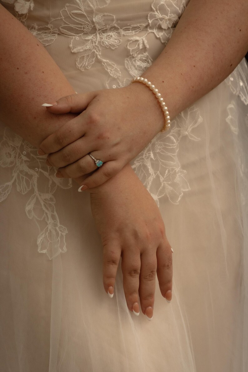 May include: A close-up of hands adorned with a pearl bracelet and a ring featuring a light blue gemstone. The nails are painted with a French manicure. The hands are resting on a white, embroidered fabric.