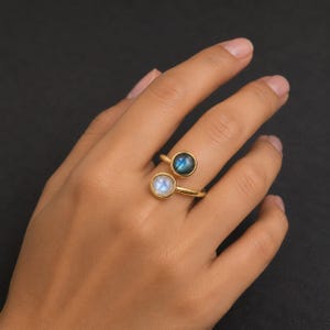 Pode incluir: Um anel aberto de tom dourado com duas pedras preciosas redondas engastadas. Uma pedra é uma pedra da lua branca translúcida e a outra é uma labradorite azul-escura com um brilho iridescente. O anel tem uma banda de metal polido.