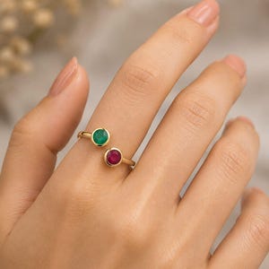 May include: A gold ring with a green emerald and a red ruby gemstone. The ring has a split shank design, with the gemstones set on opposite sides of the band. The ring is displayed on a person's finger.