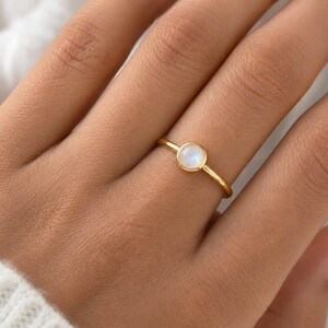 Puede incluir: Un anillo fino de oro con un pequeño cabujón de piedra de luna redondo en engaste de bisel. La piedra preciosa muestra un suave brillo blanco iridiscente, mientras que la banda es un alambre de oro pulido sencillo y continuo.