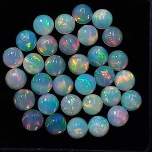 Op de afbeelding: Een verzameling ronde, in cabochon geslepen opalen gerangschikt in een cluster. Elke edelsteen heeft een doorschijnende, melkwitte basis met levendige flitsen van iriserende kleuren, waaronder groen, oranje en blauw, op de gladde oppervlakken.