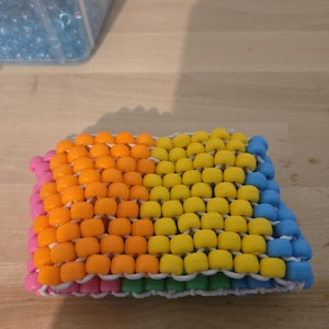 May include: A handmade beaded coaster with a rainbow color scheme. The coaster is made of small, rounded beads in shades of pink, orange, yellow, green, and blue, strung together with white thread. The coaster is rectangular in shape.