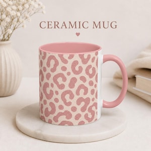 May include: A ceramic mug with a pink interior and handle. The exterior features a beige background with a pink leopard print pattern. The text "CERAMIC MUG" is displayed above the mug.