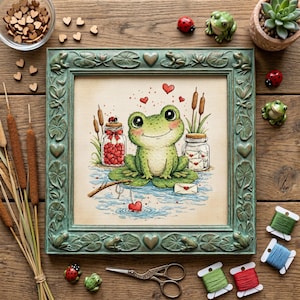 Puede incluir: Arte de punto de cruz de una rana verde sonriente en una hoja de nenúfar, pescando un corazón. La obra de arte enmarcada presenta un marco verde azulado con detalles de ranas y corazones. Frascos de corazones y cartas flanquean la rana. Objetos decorativos rodean el marco.