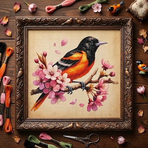 May include: Framed cross-stitch art featuring a vibrant orange and black bird perched on a flowering branch with pink blossoms. The artwork is in a decorative brown frame, surrounded by embroidery floss and small bird figurines.