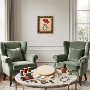Pu&ograve; includere: Un'opera d'arte incorniciata raffigura un pinguino con palloncini rossi a forma di cuore. L'opera &egrave; appesa sopra un tavolo rotondo in marmo con forniture per ricamo, tra cui rocchetti di filo, forbici e un telaio di legno. Due poltrone verdi affiancano il tavolo.