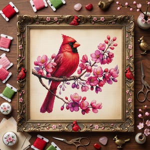 Op de afbeelding: Kruissteekkunstwerk van een felrode kardinaal op een bloeiende tak met roze bloesems. Het ingelijste stuk is omgeven door spoelen garen, decoratieve vogels en een schaar, wat wijst op een knutselthema. De lijst is versierd met bloemmotieven.