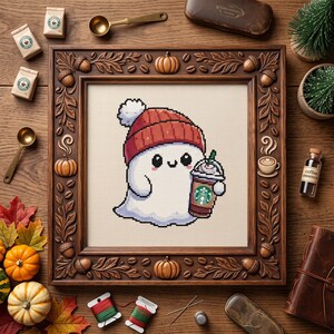 May include: A cross-stitch pattern featuring a white ghost wearing a red beanie and holding a coffee cup with a green logo, framed in a dark wood frame carved with pumpkins, acorns, and coffee beans on a wooden surface with craft supplies.
