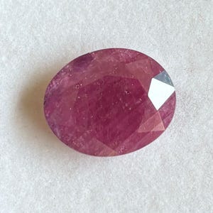 Puede incluir: Una piedra preciosa ovalada y facetada de color magenta rosado intenso. La piedra presenta una superficie pulida con múltiples facetas geométricas que reflejan la luz, creando un aspecto brillante en toda su superficie translúcida y saturada.