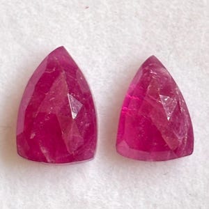 May include: Two loose, faceted gemstones with a vibrant magenta-pink hue. Each stone features a triangular, shield-like shape with multiple geometric facets that reflect light, creating a sparkling, translucent appearance on a plain white surface.
