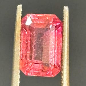 Op de afbeelding: Een rechthoekige edelsteen in smaragdgeslepen vorm met een levendige roze-rode tint en gefacetteerde oppervlakken. De steen wordt vastgehouden tussen twee metalen pincetten met gekartelde randen, wat de doorschijnende kwaliteit benadrukt.