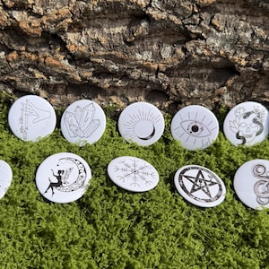 May include: A collection of white, round buttons with black line art designs. The designs include crystals, moons, eyes, a skull with a snake, a book, a fairy, a sun, a snowflake, a pentagram, and a spiral. The buttons are arranged on green moss.