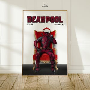 May include: A framed poster featuring Deadpool in a Santa hat, seated on a red throne. The word "DEADPOOL" is prominently displayed in large, red letters at the top. The poster has a gold frame and is set against a white wall.