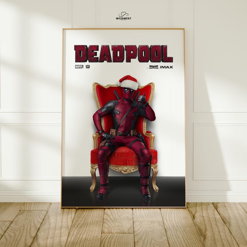 May include: A framed poster featuring Deadpool in a Santa hat, seated on a red throne. The word "DEADPOOL" is prominently displayed in large, red letters at the top. The poster has a gold frame and is set against a white wall.