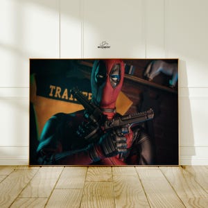 May include: Framed artwork featuring the character Deadpool in his signature red and black costume, holding two black handguns. The image has a dark, dramatic lighting scheme, with the word "TRA" visible on a yellow banner in the background.