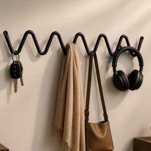May include: A black metal wall-mounted coat rack with a wavy design. Keys, a scarf, a purse, and headphones hang from the hooks. The rack is mounted on a light-colored wall, with a plant and decor in the background.
