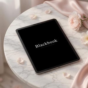 May include: A tablet device with a black screen displaying the white text Blackbook in the center. The device features a thin metallic frame and is positioned on a round surface with a white marble pattern, accompanied by light pink fabric and flowers.