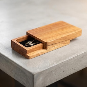 Puede incluir: Una caja de joyería de madera con tapa corredera, que muestra un collar con colgante en forma de corazón. La caja es de madera clara y descansa sobre una superficie gris. El collar tiene una cadena dorada y un colgante en forma de corazón.