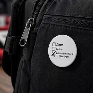 May include: A white button pinned to a black backpack. The button has the text "Single", "Taken", and "Spiritually married to John Logan" with a checkmark next to the last option. The backpack has a silver zipper.