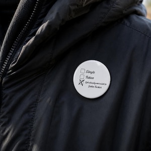Op de afbeelding: Een witte button met de tekst "Single", "Taken" en "Spiritually married to John Tucker". Het vakje "Taken" is aangekruist met een X. De button is vastgemaakt aan een zwarte jas.