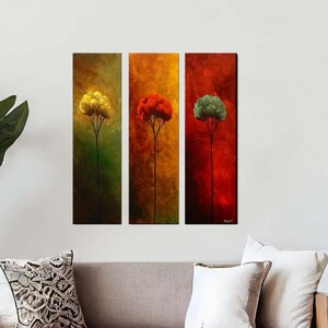 May include: Three abstract paintings of trees in different colors, yellow, red, and green, hanging on a white wall above a light brown sofa with white pillows and a coffee table with a vase of greenery.