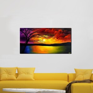 May include: A painting of a sunset over a lake with a silhouetted tree in the foreground. The sky is a vibrant mix of purple, orange, and red. The painting is framed and hanging on a white wall above a yellow sofa.