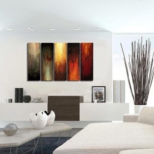 May include: Modern living room with abstract art. Five vertical panels of artwork in warm tones of gold, orange, and red hang above a white cabinet. A large white sofa with pillows and a tall plant in a white pot complete the scene.