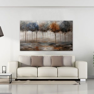 May include: A painting of a forest scene with trees in shades of blue, brown, and green. The trees are silhouetted against a misty background. The painting is displayed on a white wall above a white sofa with brown pillows.