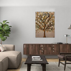 May include: A painting of a gold tree with a textured, abstract style. The tree is set against a light brown background. The painting is framed and hanging on a white wall in a living room setting.